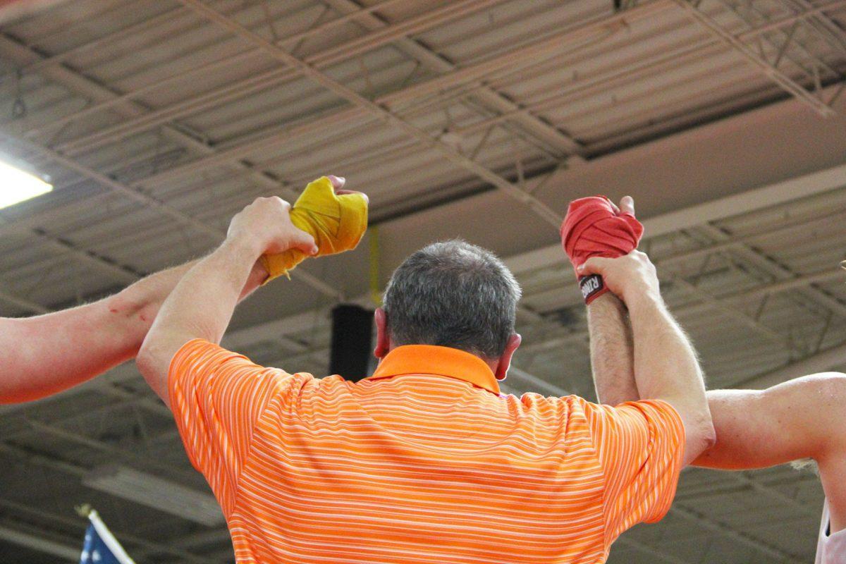 A referee holds the arms of two boxers after their match, waiting for the emcee to announce the winner.