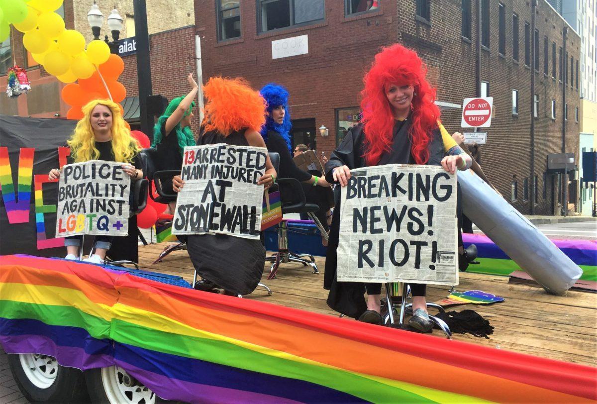 Three women wearing brightly colored wigs hold up posters resembling newspaper headlines. 