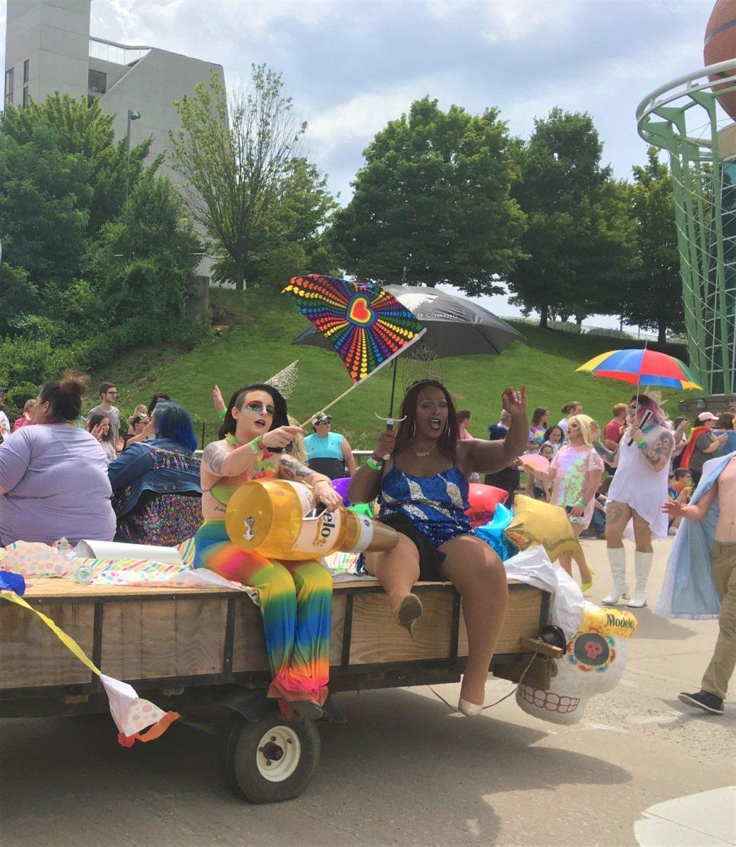Dressed in colorful outfits, makeup, and hair, two people wave at the crowd. 